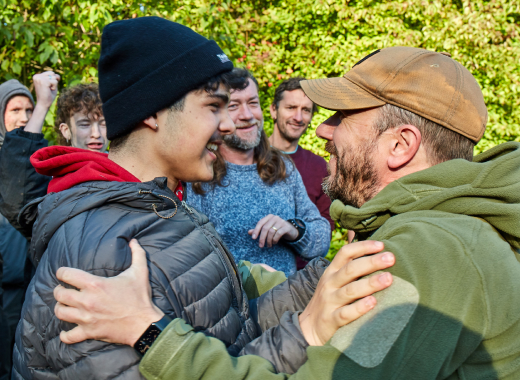A mentor in a cap embracing a smiling boy, with other boys and men around them