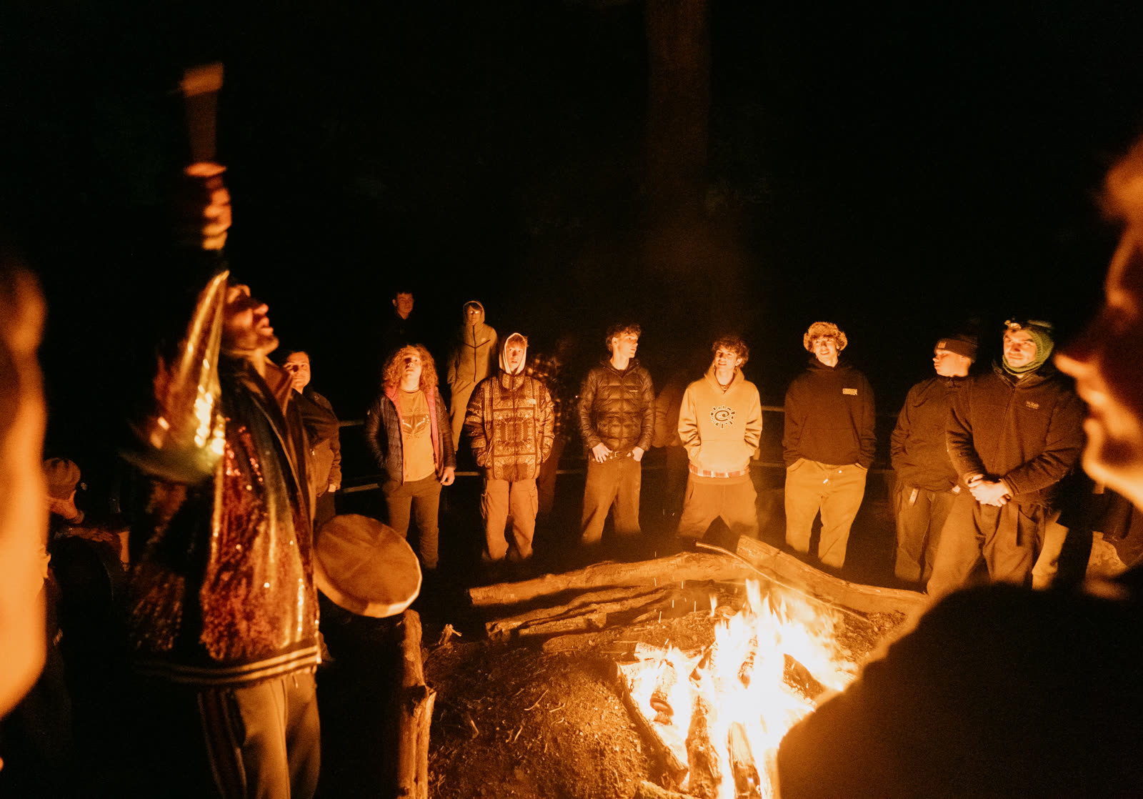 Boys and men standing in a circle around the fire at night, a mentor with a drum raising a torch