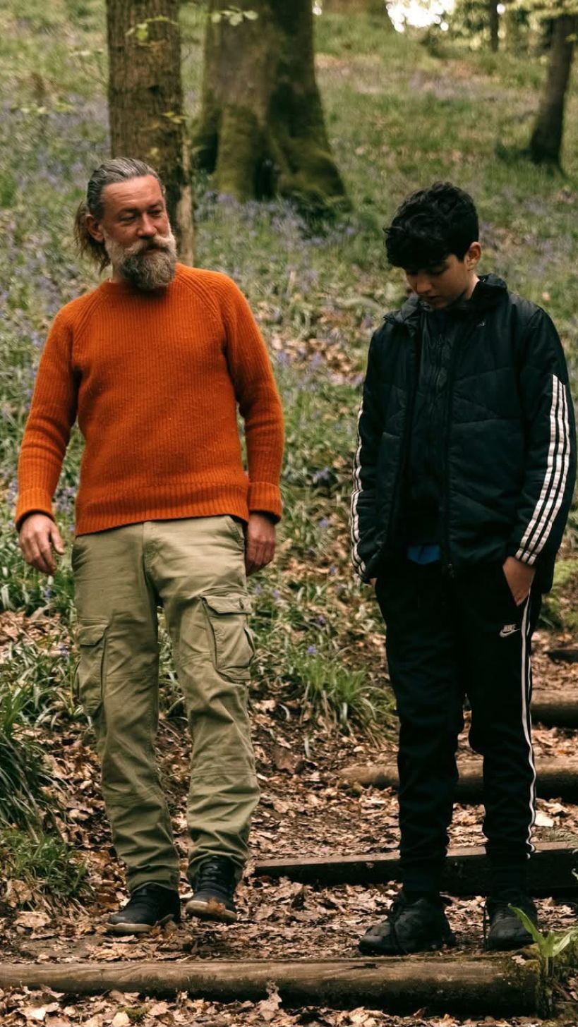 A bearded mentor in an orange jumper walking with a boy through bluebell woods